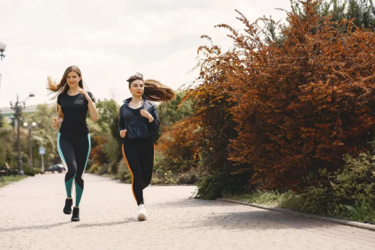 Arlington VA 19 Two young women jogging outdoors on a paved path, surrounded by greenery and autumn-colored bushes.