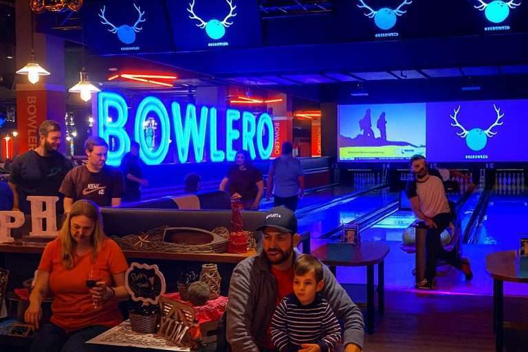 Arlington VA 15 People bowling in a vibrant alley with neon "BOWLERO" sign, screens, and party decorations; a man and child in foreground.