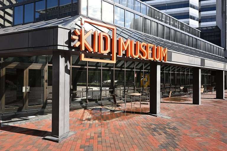 Arlington VA 18 KID Museum entrance with orange sign, glass façade, and brick pavement in a modern building complex.