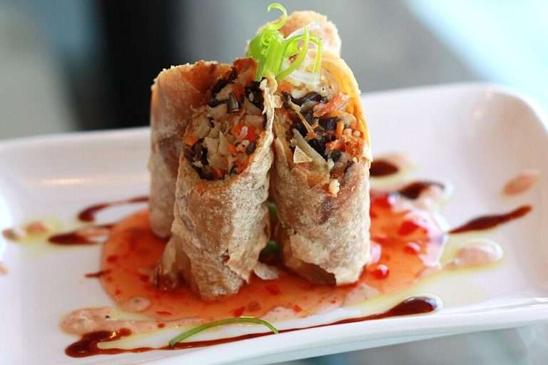 Arlington VA 35 Three crispy spring rolls on a white plate, garnished with green onions and drizzled with sweet chili sauce.