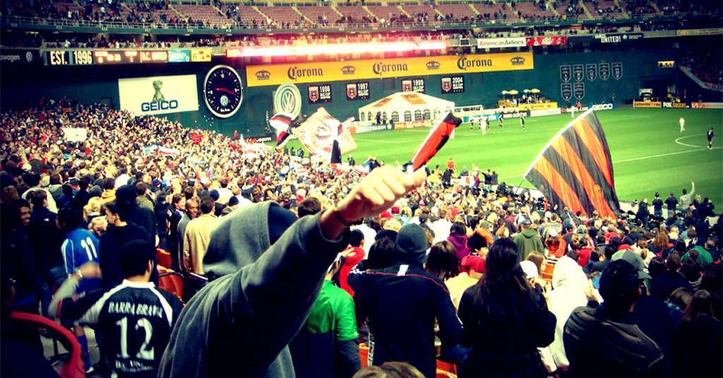 A large crowd of fans, many waving flags and banners, fills the stands at RFK Stadium during a DC United soccer match, with the pitch and players visible in the background.