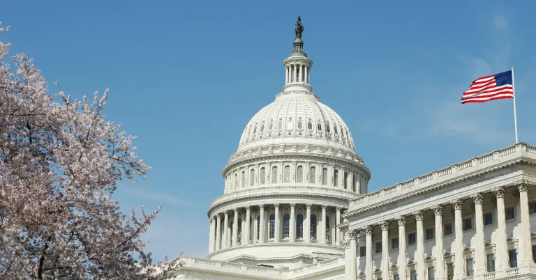 The U.S. Capitol dome in Washington, D.C., with an American flag waving and cherry blossom trees in bloom, symbolizing government, politics, and national heritage.