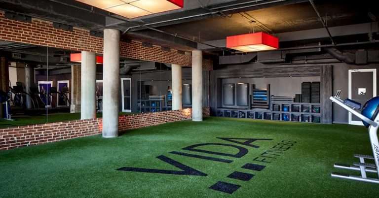Interior view of VIDA Fitness U Street location, featuring a spacious training area with artificial turf, mirrored walls, exposed brick and concrete pillars, red accent lighting, and organized fitness equipment along the back wall.