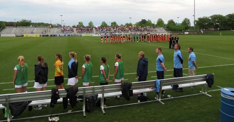 Washington Spirit players and coaching staff line up on the sideline before an NWSL match, with both teams lined up on the field and fans in the stands in the background.