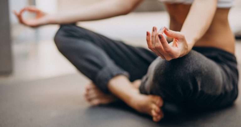 Close-up of a person sitting cross-legged on a yoga mat, meditating with hands resting on knees in a mudra position, wearing casual yoga attire in a serene indoor studio setting.